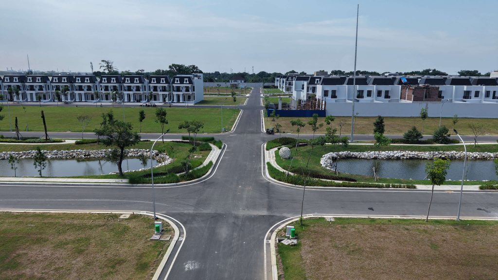 Aerial view of a modern housing development with white townhouses, wide intersecting roads, and a small pond surrounded by landscaped greens.
