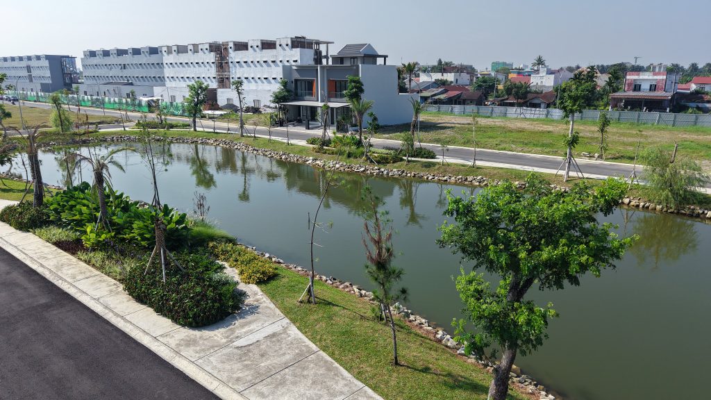 Wide view of a canal with a paved path and newly planted trees, a modern two-story building under construction along the bank, and distant houses.
