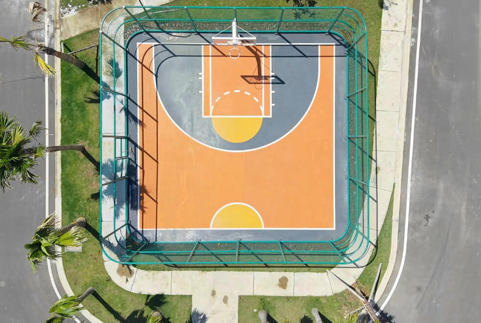 Aerial view of an outdoor basketball court inside a teal fence, with an orange playing surface, white boundary lines, and a yellow center circle; a hoop is at the far end. Nearby palm trees and a street frame the scene.